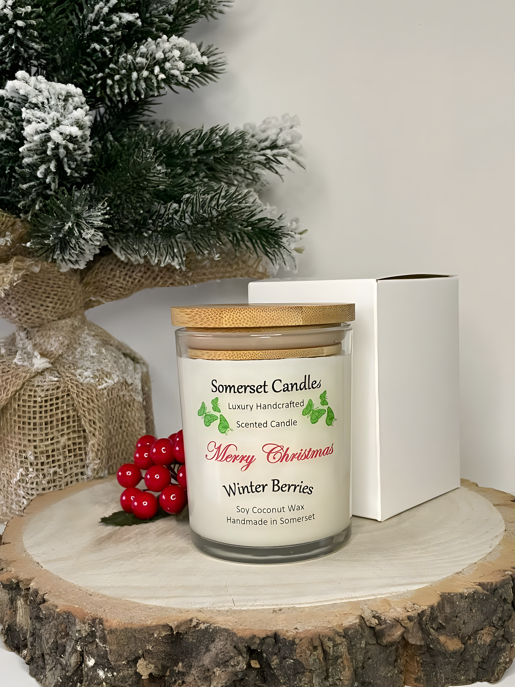 Christmas Winter Berries soy candle is displayed on a wooden board with berries, a small Christmas tree and a white gift box behind. The candle is white in colour with bright green butterflies above the scent name and has a bamboo lid fitted.