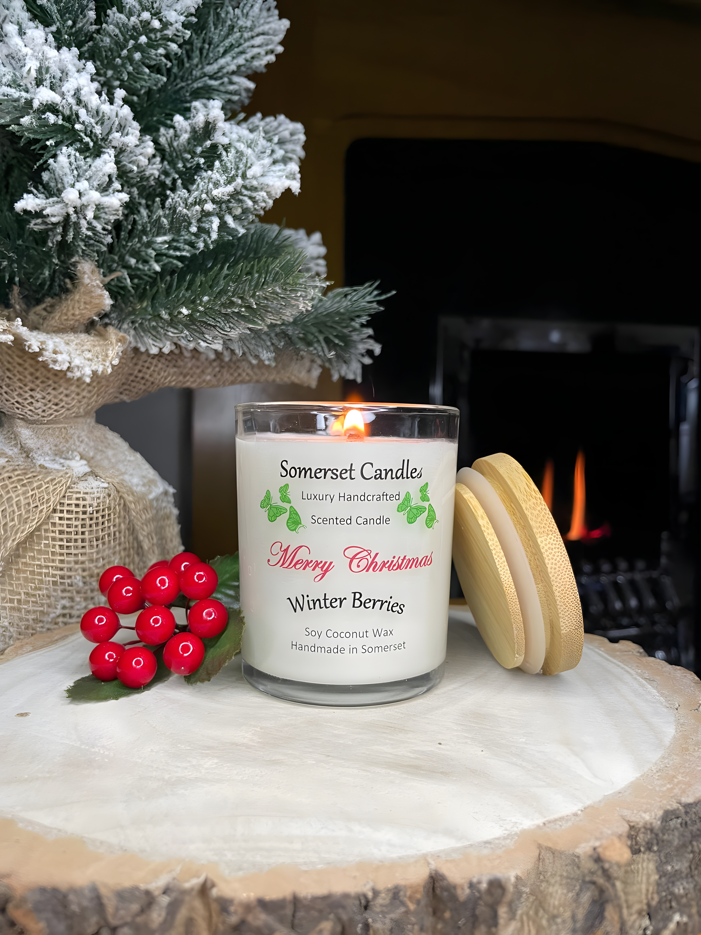 Christmas Winter Berries soy candle is displayed on a wooden board with berries behind, in front of a lit fireplace. The candle is white in colour with bright green butterflies above the scent name and has a bamboo lid resting up against it.