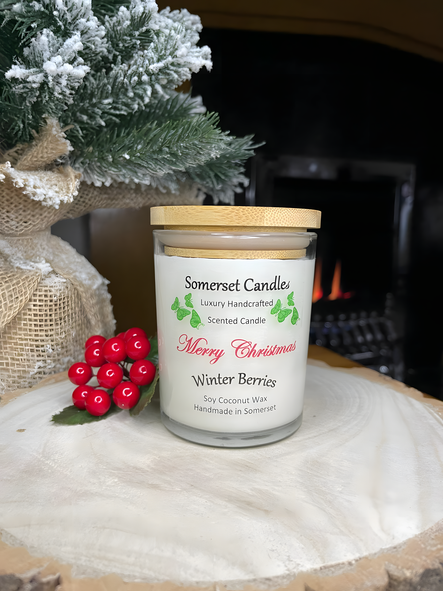 Christmas Winter Berries soy candle is displayed on a wooden board in front of a lit fire with berries and a small Christmas tree behind. The candle is white in colour with bright green butterflies above the scent name and has a bamboo lid is fitted.