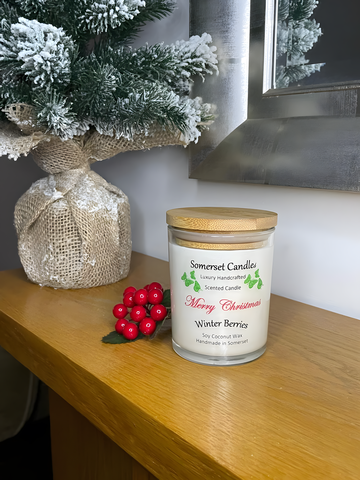 Christmas Winter Berries soy candle is displayed on a wooden mantel piece with berries and a small Christmas tree behind. The candle is white in colour with bright green butterflies above the scent name and has a bamboo lid fitted.