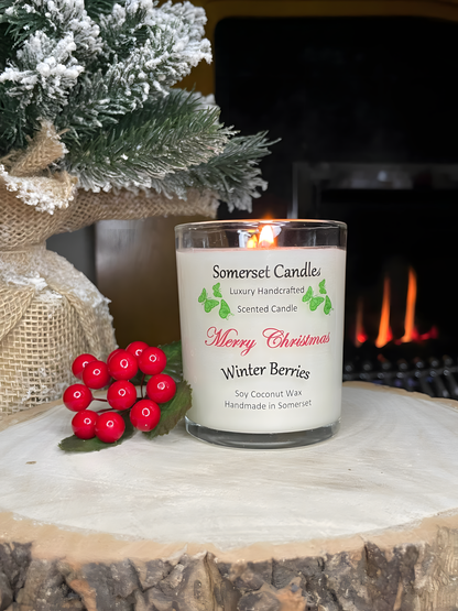 Merry Christmas Winter Berries soy candle is displayed on a wooden board in front of a lit fire with berries and a Christmas tree to the left. The candle is white in colour with bright green butterflies above the scent name and the candle is lit.