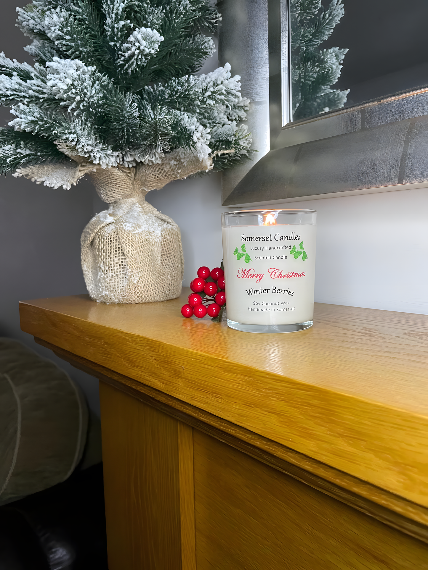 Christmas Winter Berries soy candle is displayed on a wooden mantel piece with berries and a small Christmas tree behind. The candle is white in colour with bright green butterflies above the scent name and the candle is lit.