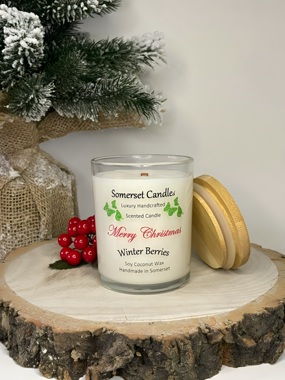 Christmas Winter Berries soy candle is displayed on a wooden board with berries and a small Christmas tree behind. The candle is white in colour with bright green butterflies above the scent name and has a bamboo lid resting up against it.