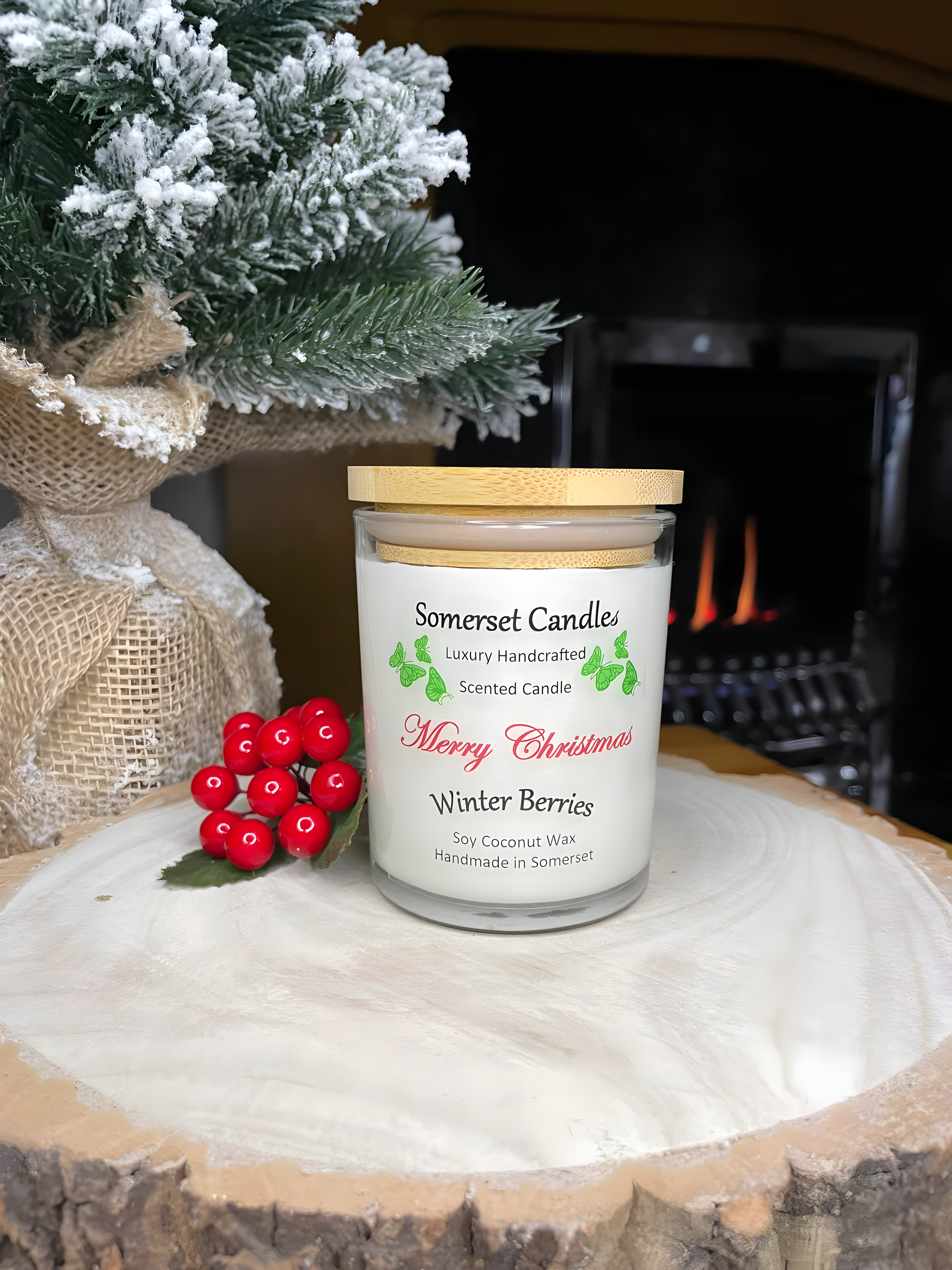 Christmas Winter Berries soy candle is displayed on a wooden board with berries, a small Christmas tree behind and is in front of a lit fire. The candle is white in colour with bright green butterflies above the scent name and has a bamboo lid fitted