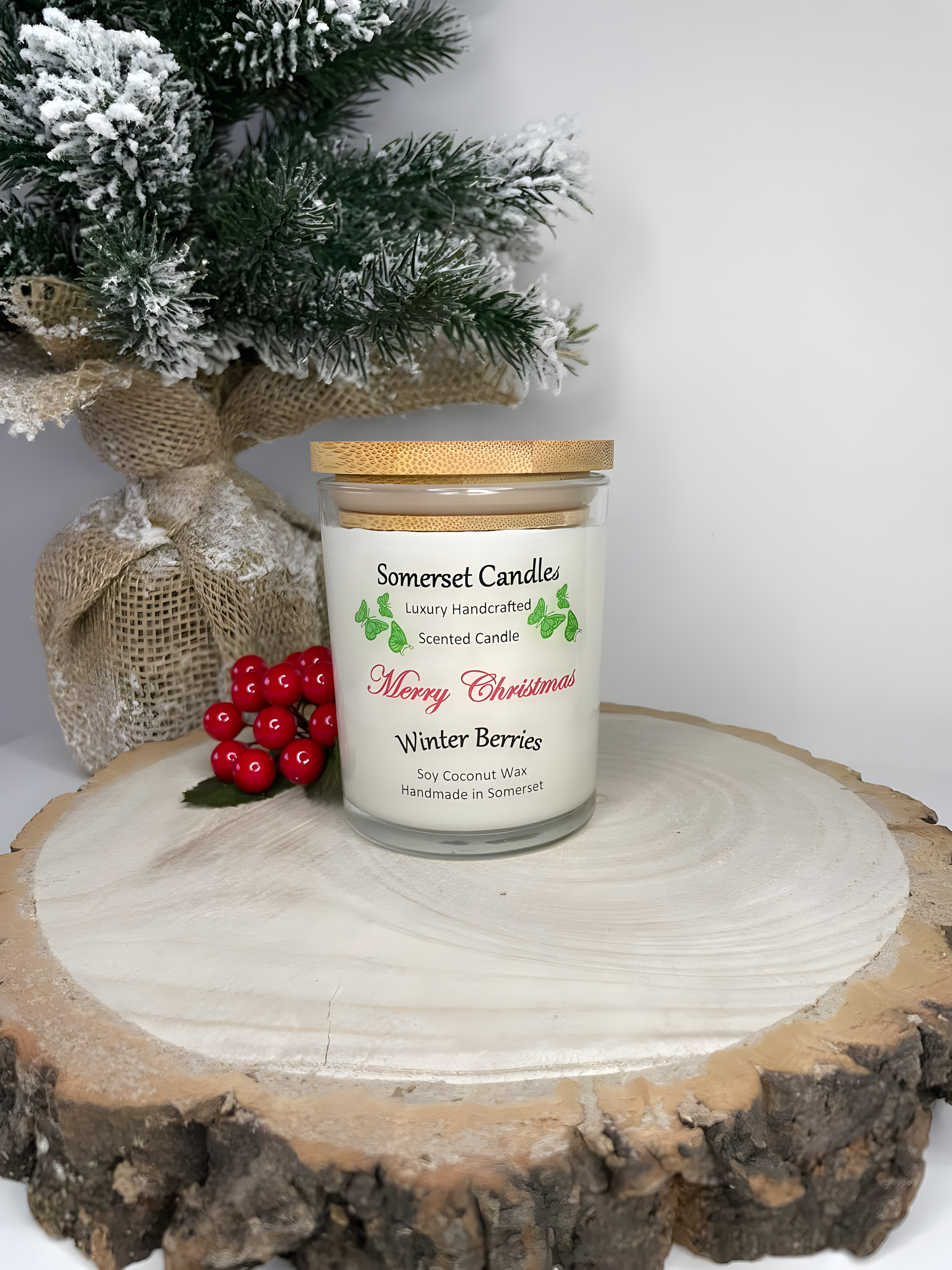 Christmas Winter Berries soy candle is displayed on a wooden board with berries and a small Christmas tree to the left behind it. The candle is white in colour with bright green butterflies above the scent name and has a bamboo lid is fitted.