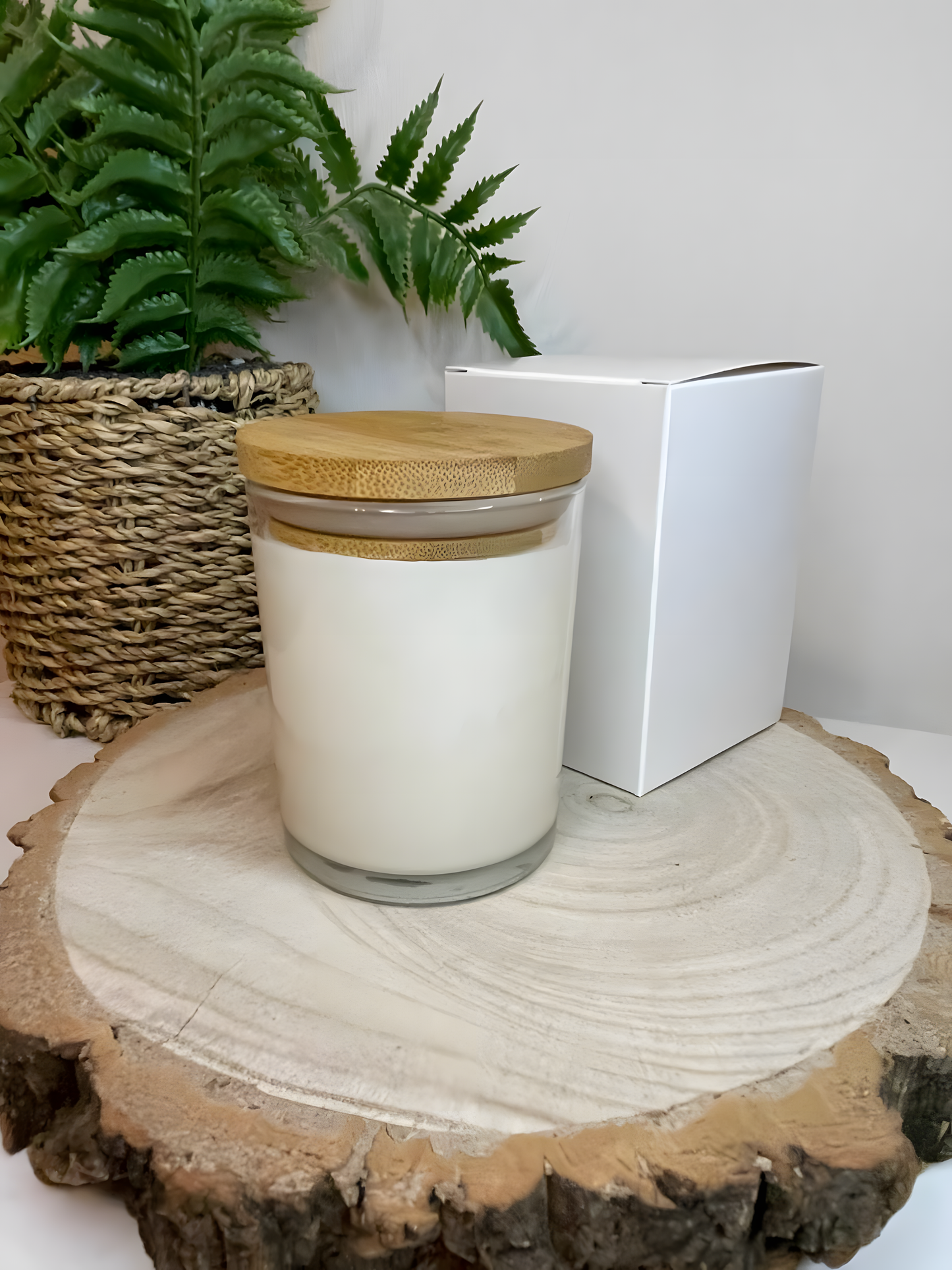 No label scented soy candle displayed on a wooden board with a white gift box and lovely plant behind. The candle is white in colour with a bamboo lid. The candle is in a clear glass jar.