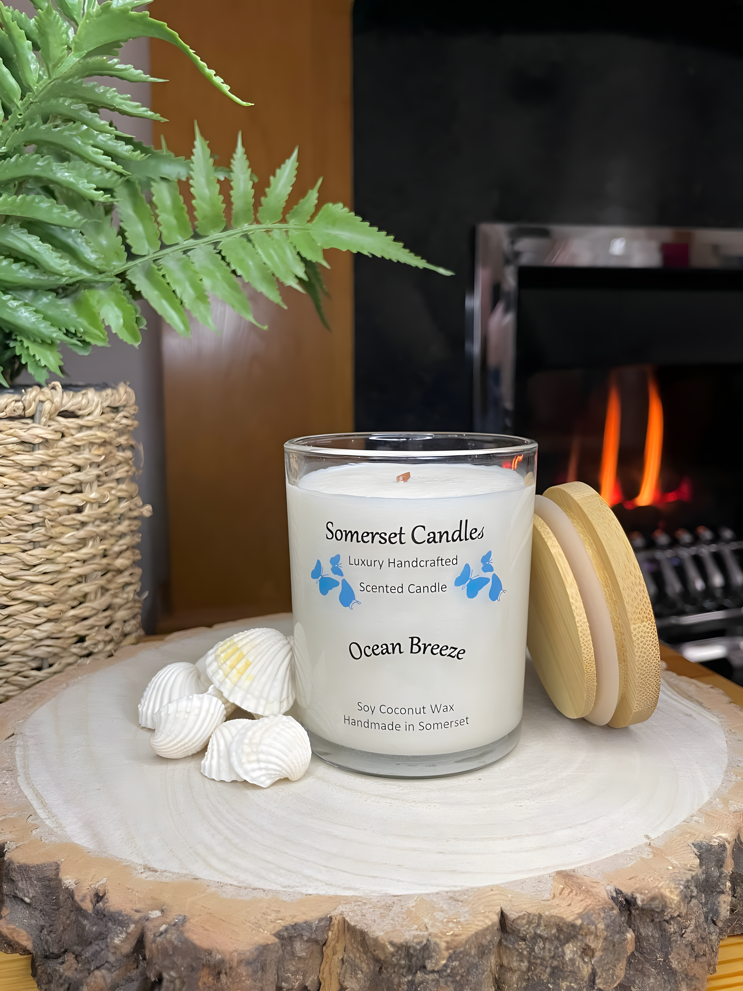 Ocean Breeze scented soy candle displayed on a wooden board with beautiful white shells in front of a lit fire. The candle is white in colour with bright blue butterflies fluttering above the scent name. A bamboo lid is lent against the candle.