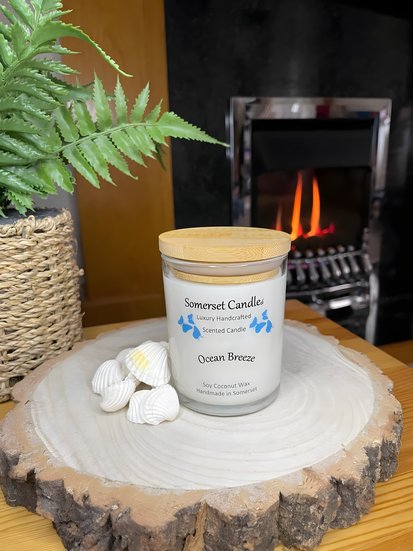 Ocean Breeze scented soy candle displayed on a wooden board with beautiful white shells in front of a lit fire. The candle is white in colour with bright blue butterflies fluttering above the scent name and fitted with a lovely bamboo lid.