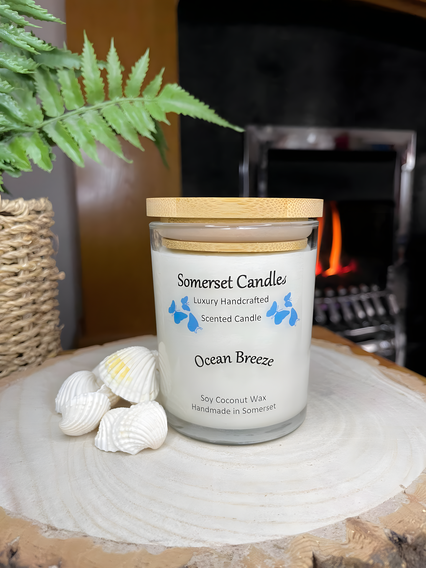 Ocean Breeze scented soy candle displayed on a wooden board with beautiful white shells and a lit fire in the back ground. The candle is white in colour with bright blue butterflies fluttering above the scent name and fitted with a lovely bamboo lid.