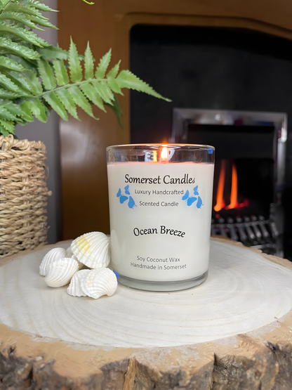 Ocean Breeze scented soy candle displayed on a wooden board with beautiful white shells in front of a lit fire. The candle is white in colour with bright blue butterflies fluttering above the scent name. The candle is lit.