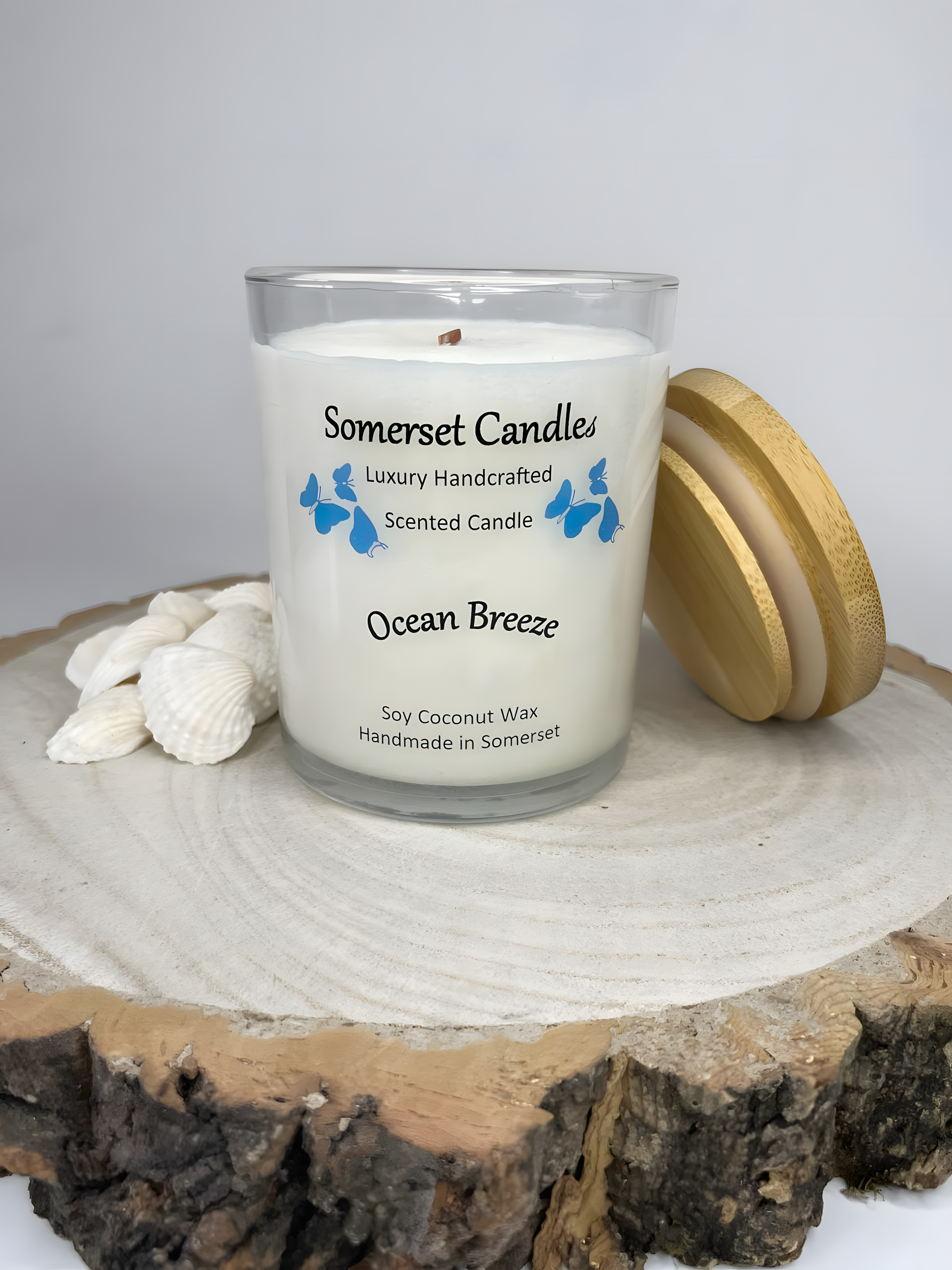 Ocean Breeze  scented soy candle displayed on a wooden board with beautiful white shells. The candle is white in colour with bright blue butterflies fluttering above the scent name and has a bamboo lid lent against the right side of the candle.