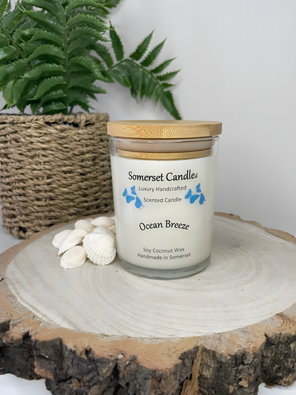 Ocean Breeze Scented soy candle  displayed on a wooden board with beautiful white shells. The candle is white in colour with bright blue butterflies fluttering above the scent name and fitted with a lovely bamboo lid.