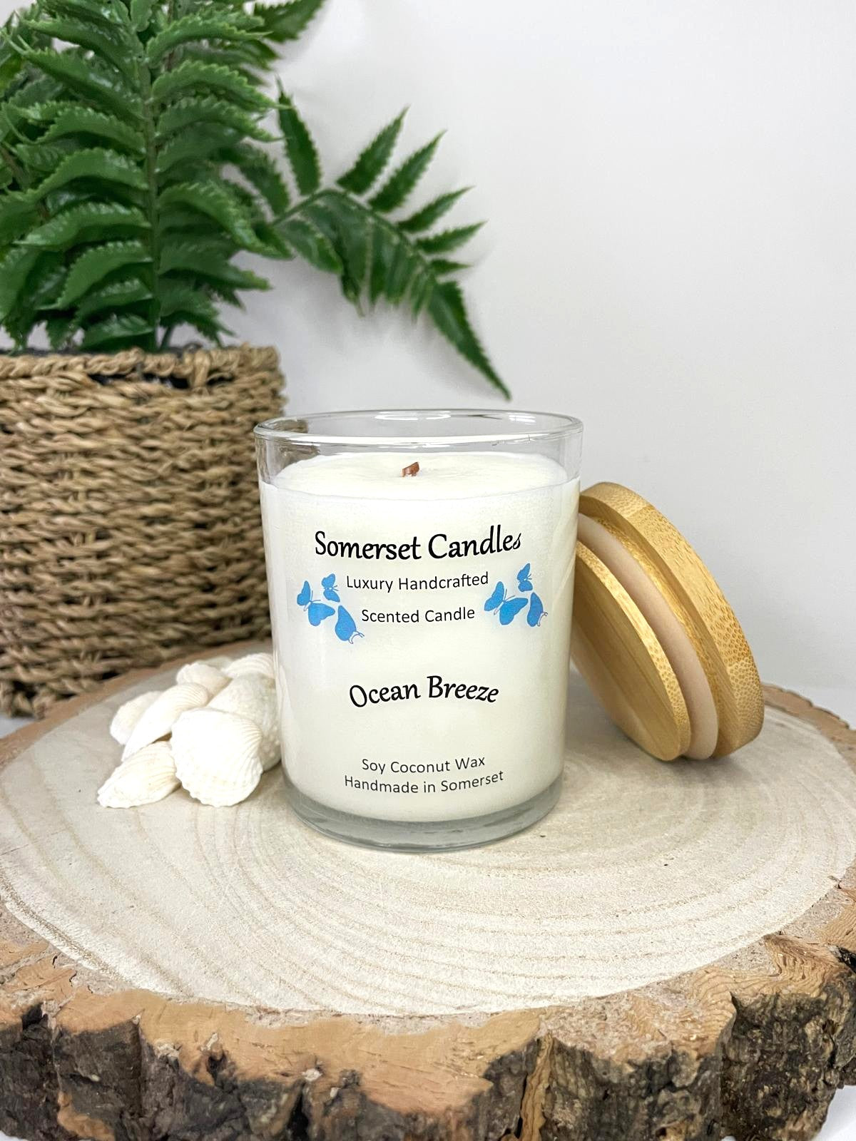White soy candle in a 7oz clear glass jar with a natural wooden lid. The candle is made from a soy-coconut blend and scented with a fresh scent of ocean breeze. Above the scent name on the label, several blue butterflies are displayed, adding a romantic touch. Clean, minimalist design ideal for gifts, home decor or self-care.