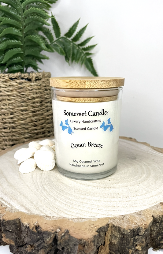 White soy candle in a 7oz clear glass jar with a natural wooden lid. The candle is made from a soy-coconut blend and scented with a fresh scent of ocean breeze. Above the scent name on the label, several blue butterflies are displayed, adding a romantic touch. Clean, minimalist design ideal for gifts, home decor or self-care.