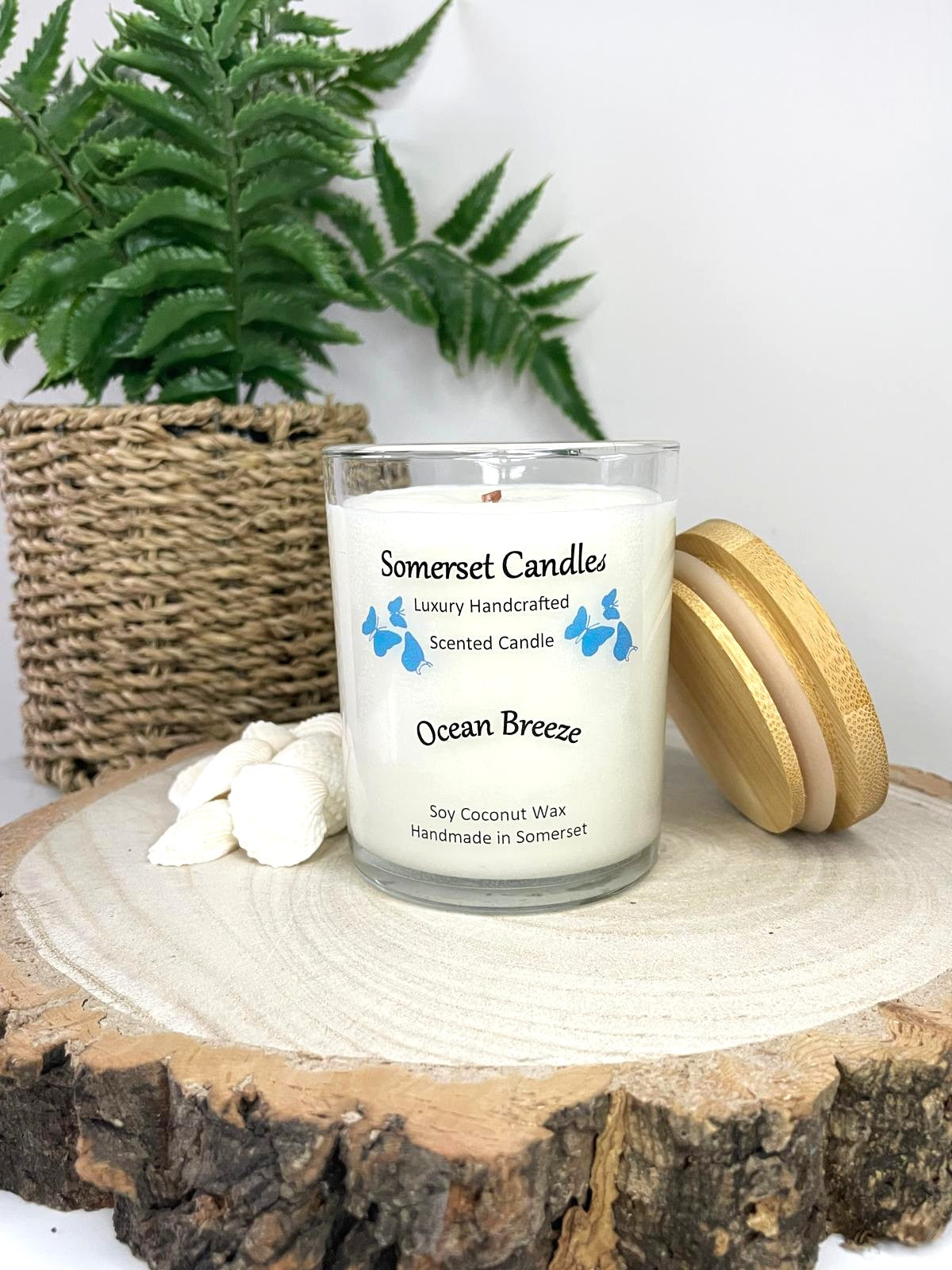 White soy candle in a 7oz clear glass jar with a natural wooden lid. The candle is made from a soy-coconut blend and scented with a fresh scent of ocean breeze. Above the scent name on the label, several blue butterflies are displayed, adding a romantic touch. Clean, minimalist design ideal for gifts, home decor or self-care.