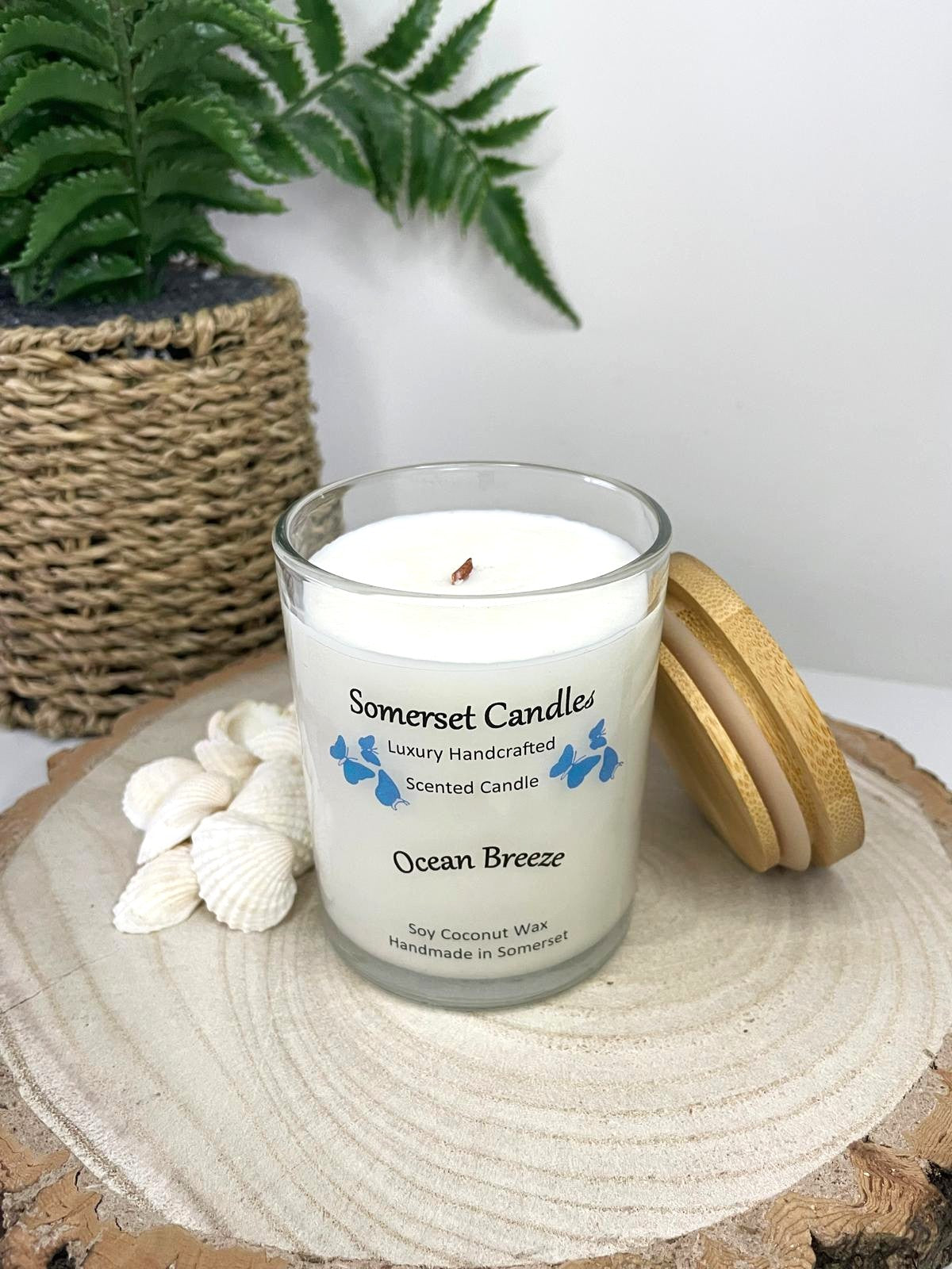 White soy candle in a 7oz clear glass jar with a natural wooden lid. The candle is made from a soy-coconut blend and scented with a fresh scent of ocean breeze. Above the scent name on the label, several blue butterflies are displayed, adding a romantic touch. Clean, minimalist design ideal for gifts, home decor or self-care.