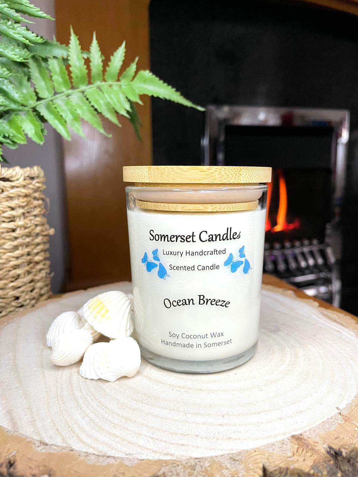 White soy candle in a 7oz clear glass jar with a natural wooden lid. The candle is made from a soy-coconut blend and scented with a fresh scent of ocean breeze. Above the scent name on the label, several blue butterflies are displayed, adding a romantic touch. Clean, minimalist design ideal for gifts, home decor or self-care.