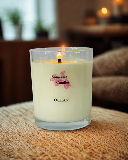 A lit Somerset Candles 'Ocean' scented candle in a clear glass jar, resting on a textured beige woven blanket with a cozy, warm-toned background.