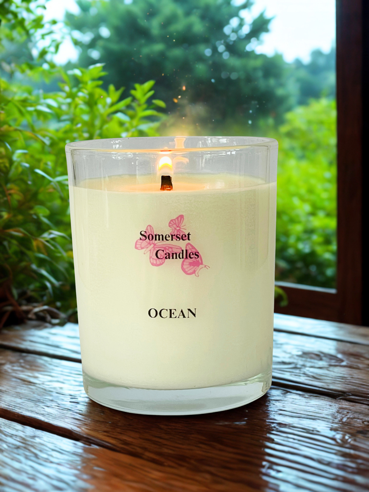 A lit white coconut wax candle in a clear glass jar sits on a wooden table outdoors, with green foliage visible in the background. The label reads 'Somerset Candles' and 'OCEAN' with pink butterfly graphics.