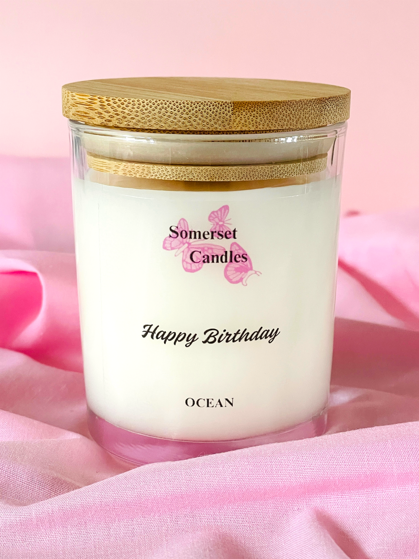 Somerset Candles 'Happy Birthday Ocean' candle with a wooden lid on a pink fabric background
