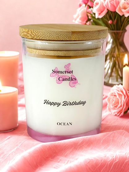 Somerset Candles 'Happy Birthday Ocean' candle on a pink fabric background with flowers and another candle.