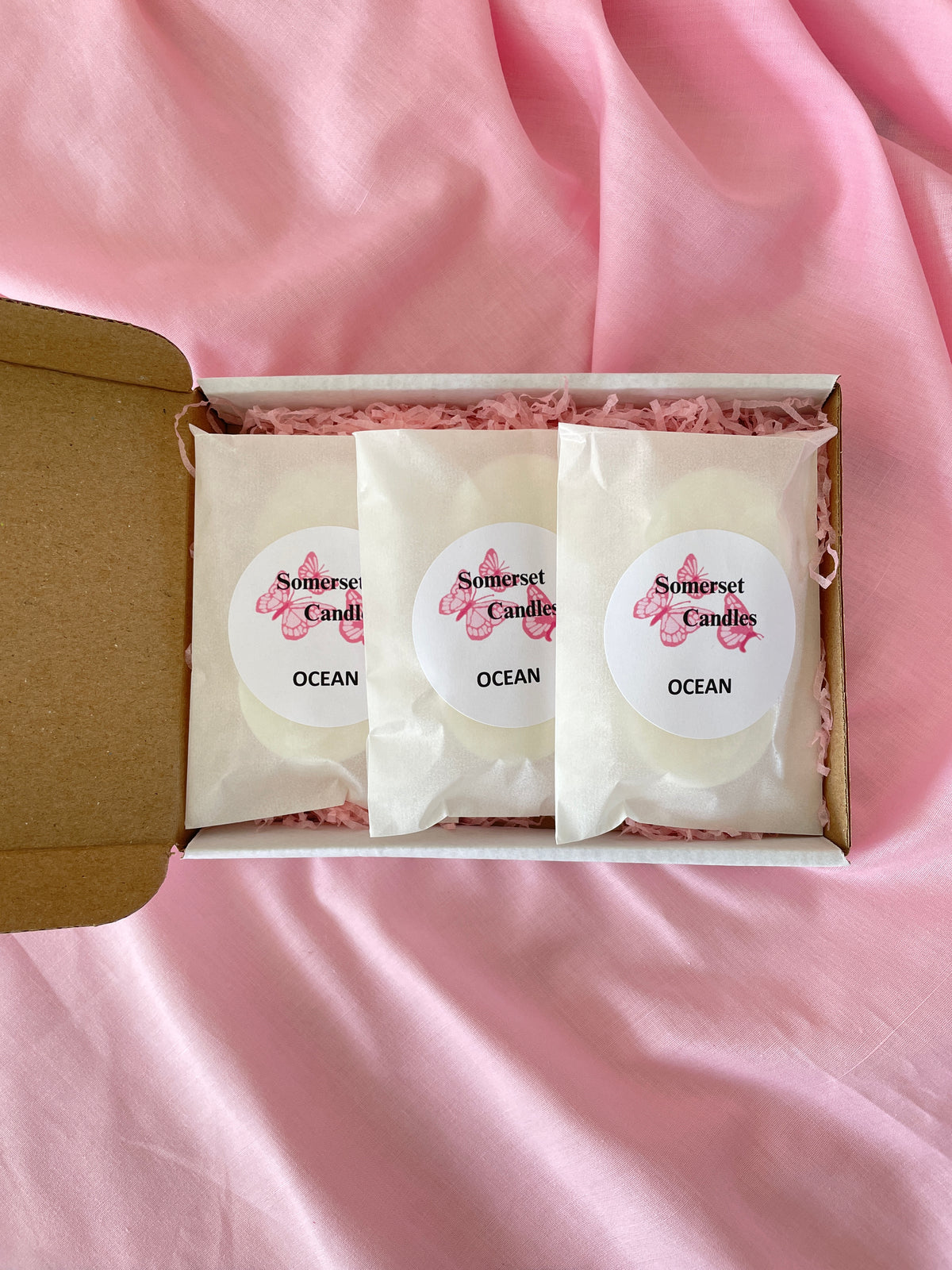Three individually wrapped Somerset Candles wax melts 'Ocean' scented wax melts are displayed in a white cardboard gift box lined with pink shredded paper, set against a draped pink fabric background.