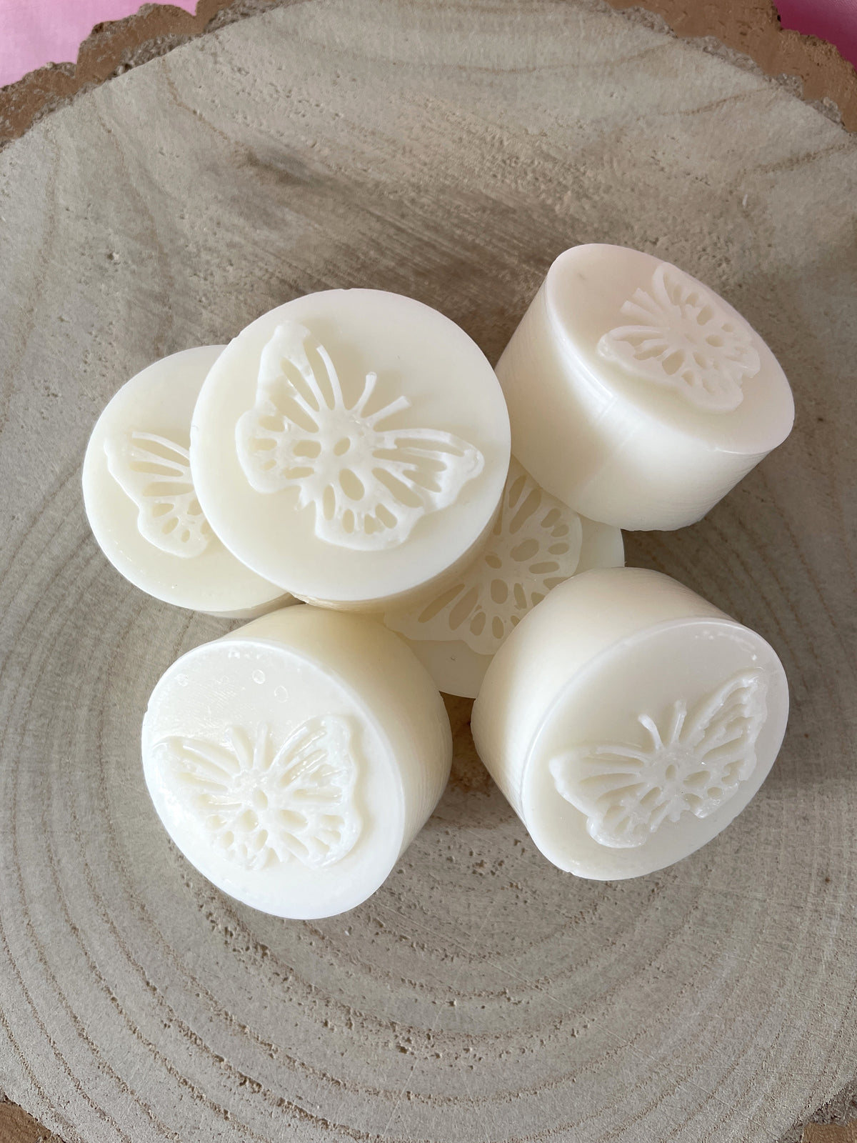 Six round white wax melts with embossed butterfly designs, arranged on a rustic wood slice background.