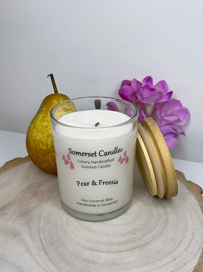 Pear & Freesia Scented soy candle displayed on a wooden board with a pear & a freesia behind. The candle is white in colour with bright pink butterflies above the scent name with a lovely bamboo lid lent against the right side of the candle.