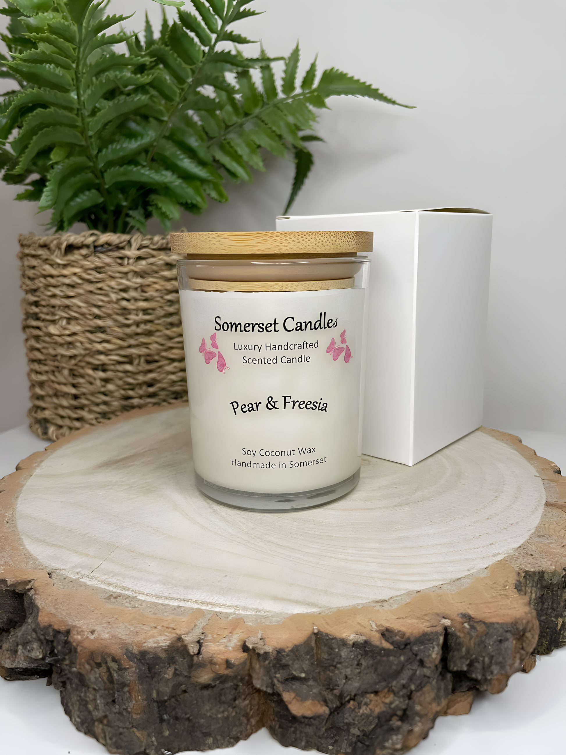 Pear & Freesia scented soy candle displayed on a wooden board with a Gift box and a plant behind. The candle is white in colour with bright pink butterflies above the scent name with a lovely bamboo lid fitted.