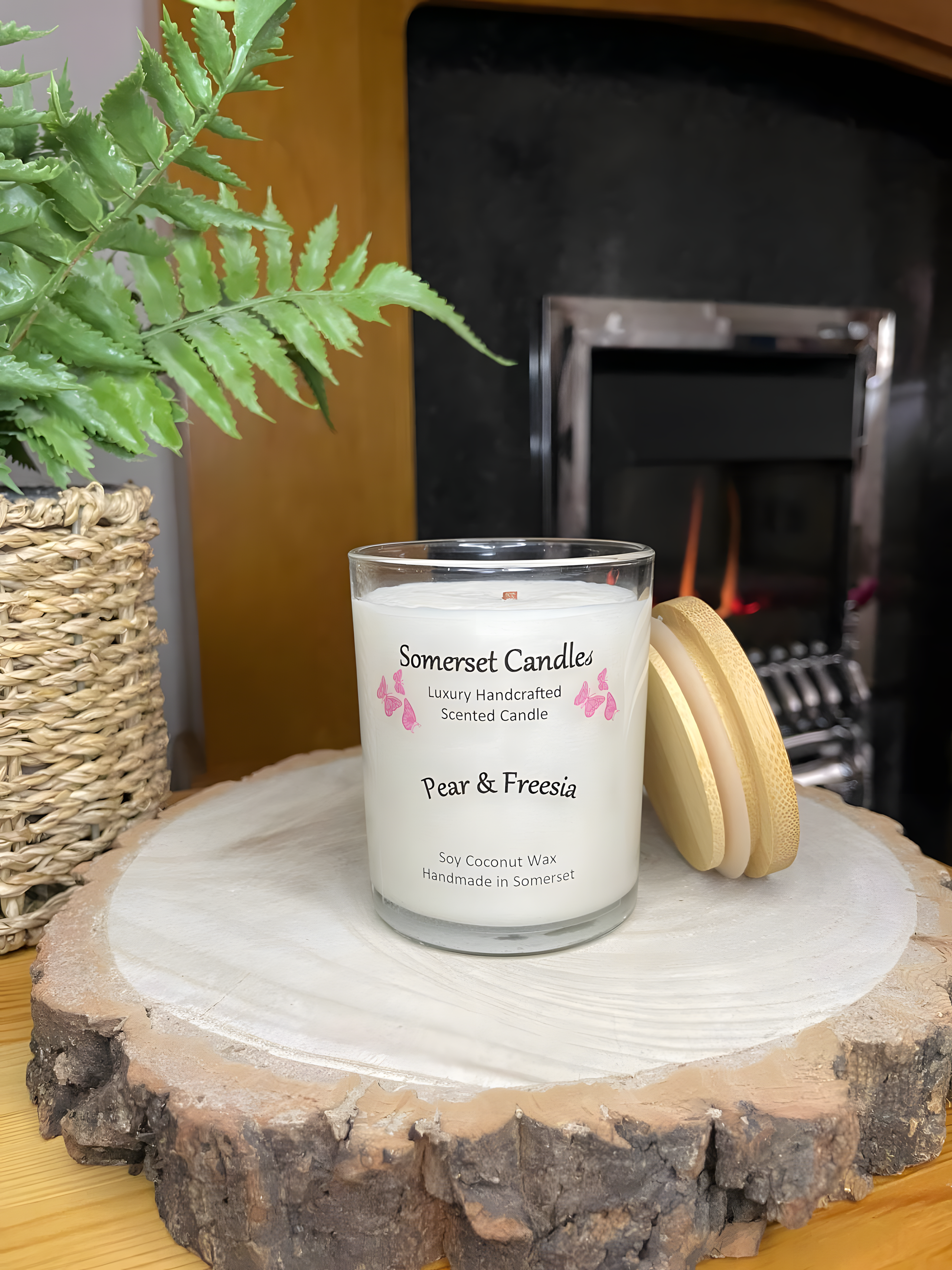 Pear & Freesia scented soy candle displayed on a wooden board with plant and a lit fire behind. The candle is white in colour with bright pink butterflies above the scent name with a lovely bamboo lid lent against the right side of the candle.