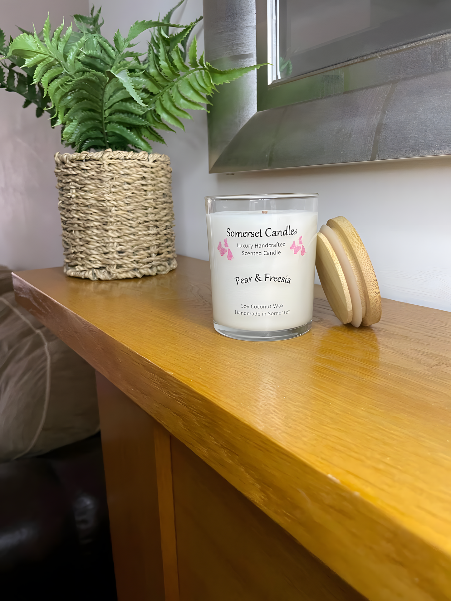 Pear & Freesia scented soy candle displayed on a wooden mantel piece with a plant behind. The candle is white in colour with bright pink butterflies above the scent name with a lovely bamboo lid lent against the right side of the candle.