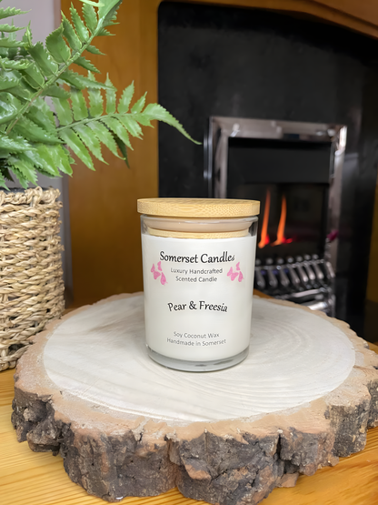 Pear & Freesia scented soy candle displayed on a wooden board with plant and a lit fire behind. The candle is white in colour with bright pink butterflies above the scent name with a lovely bamboo lid fitted.