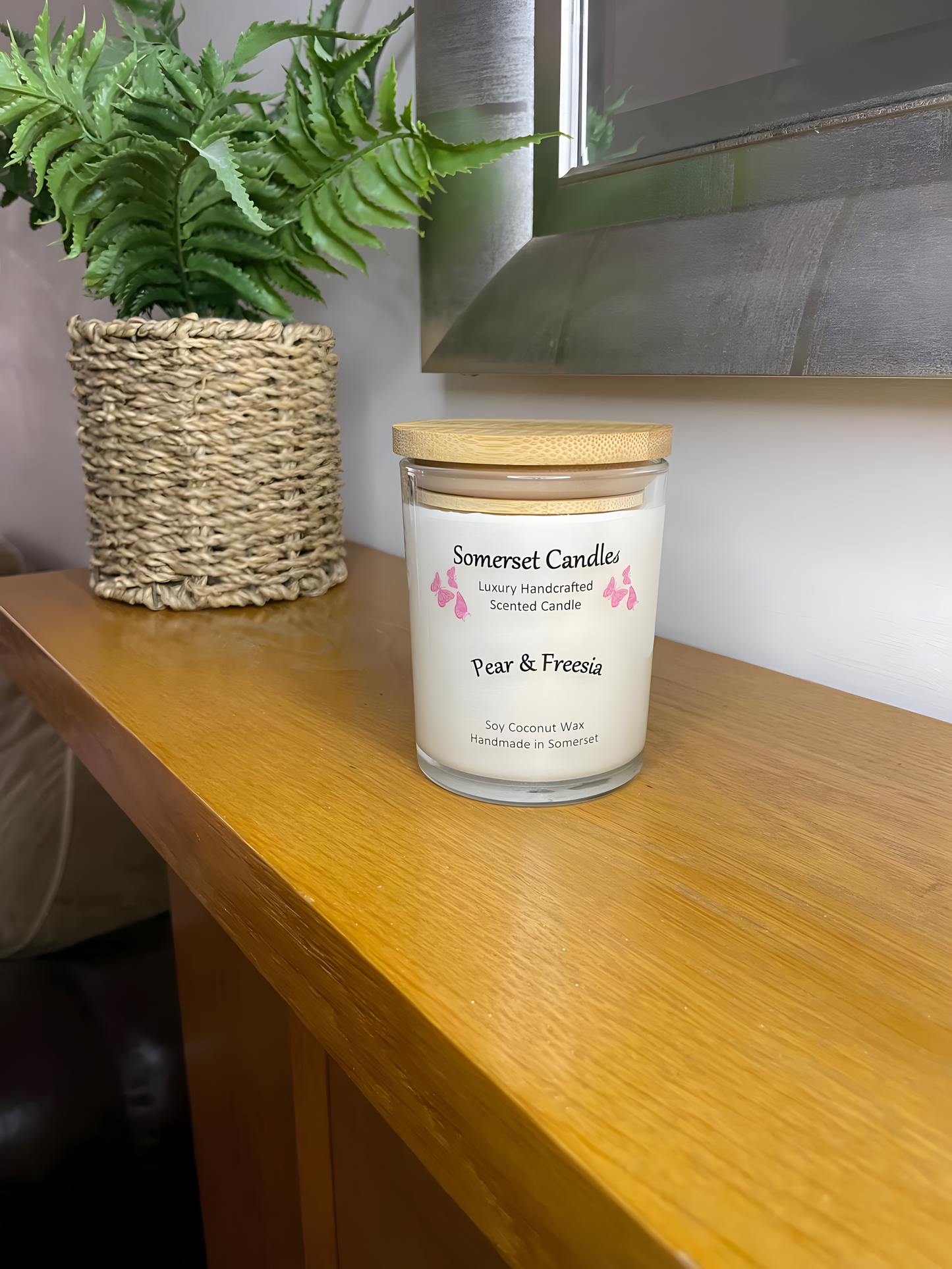 Pear & Freesia scented soy candle displayed on a wooden mantel piece with a plant behind. The candle is white in colour with bright pink butterflies above the scent name with a lovely bamboo lid fitted to the clear glass container.