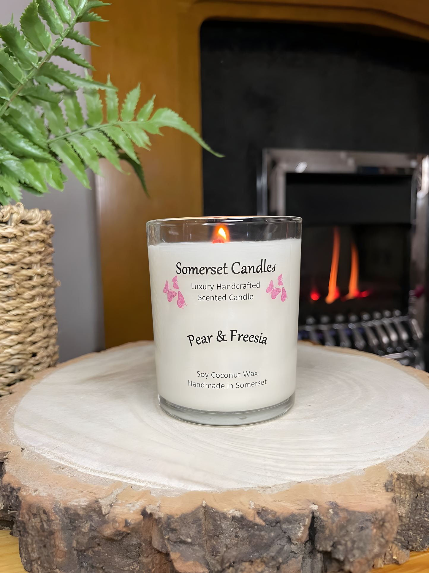 Pear & Freesia scented soy candle displayed on a wooden board in front of a lit fire. The candle is white in colour with bright pink butterflies above the scent name. The candle is lit and the is a plant to the left of the candle.