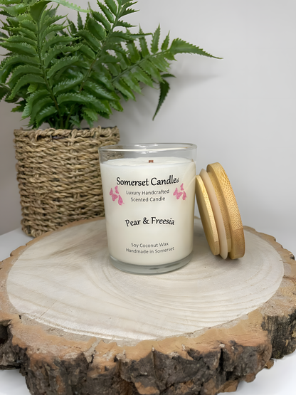 Pear & Freesia scented soy candle displayed on a wooden board with a plant behind. The candle is white in colour with bright pink butterflies above the scent name with a lovely bamboo lid lent against the right side of the candle.