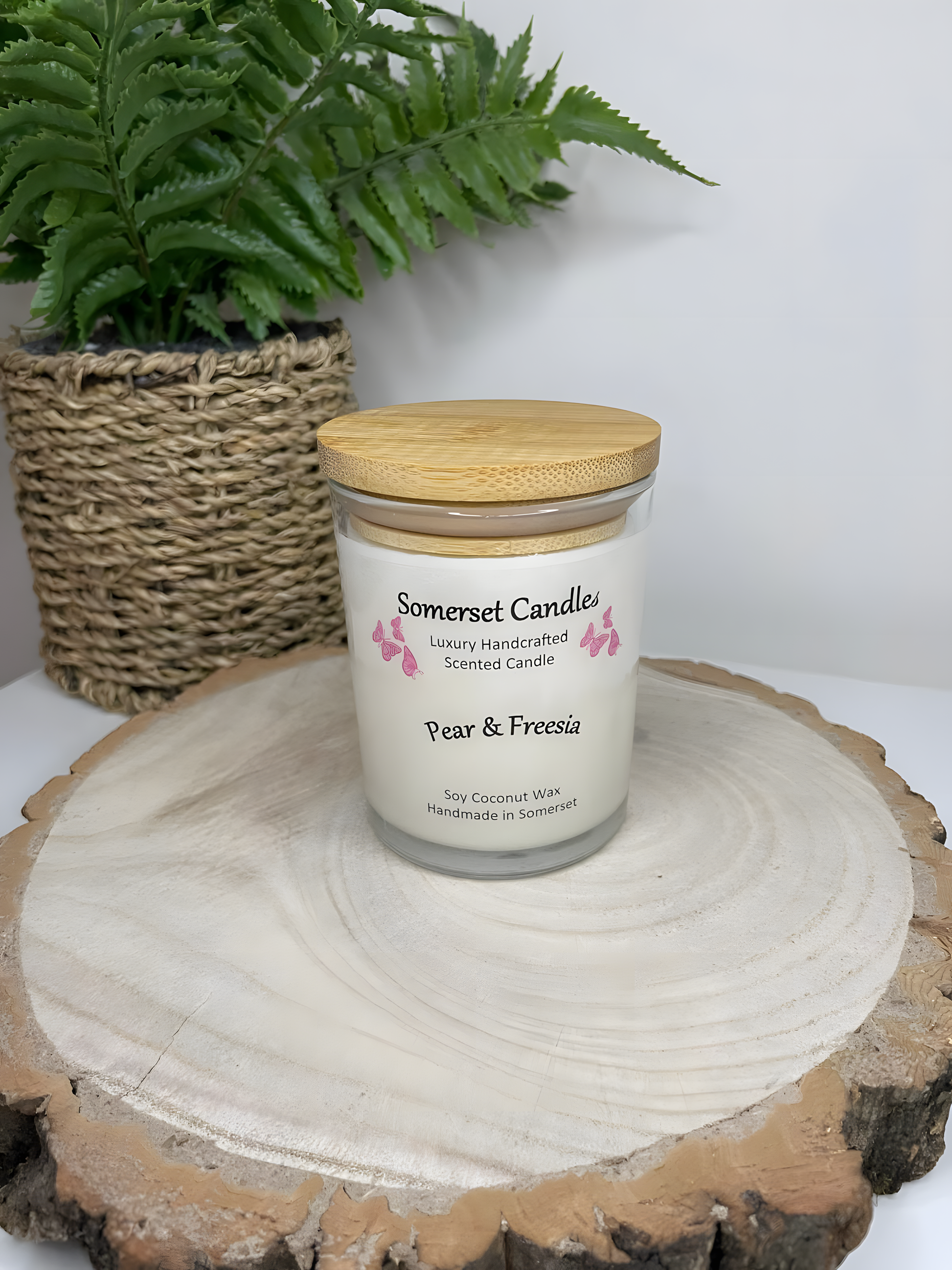 Pear & Freesia scented soy candle displayed on a wooden board. The candle is white in colour with bright pink butterflies above the scent name with a lovely bamboo lid fitted to the glass candle container.