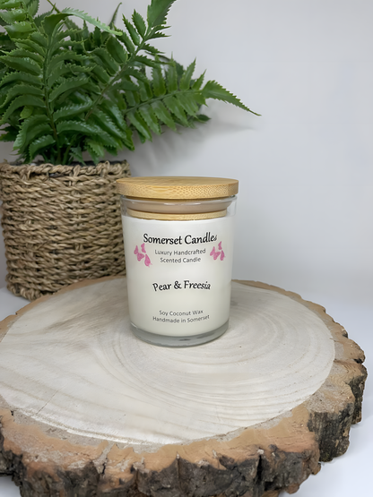 Pear & Freesia scented soy candle displayed on a wooden board with a plant behind. The candle is white in colour with bright pink butterflies above the scent name with a lovely bamboo lid fitted.