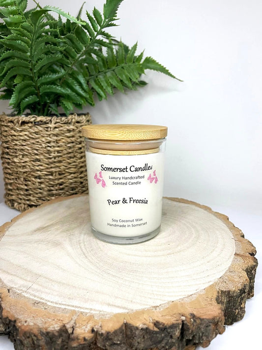 White soy candle in a 7oz clear glass jar with a natural wooden lid. The candle is made from a soy-coconut blend and scented with a delicate mix of pear and freesia. Above the scent name on the label, several pink butterflies are displayed, adding a romantic touch. Clean, minimalist design ideal for gifts, home decor or self-care.
