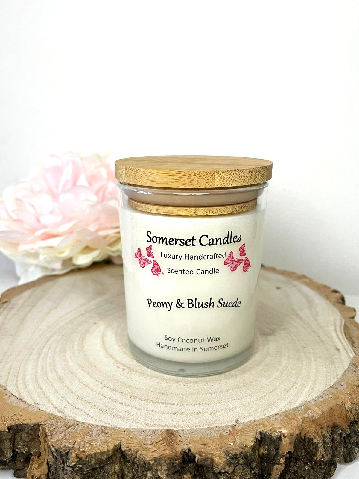 White soy candle in a 7oz clear glass jar with a natural wooden lid. The candle is made from a soy-coconut blend and scented with a soft elegant mix of Peony and Blush Suede. Above the scent name on the label, several pink butterflies are displayed, adding a romantic touch. Clean, minimalist design ideal for gifts, home decor or self-care.