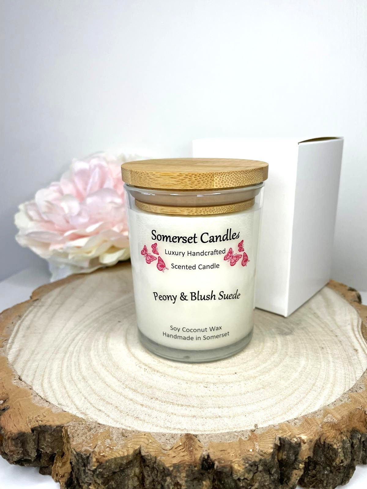 White soy candle in a 7oz clear glass jar with a natural wooden lid. The candle is made from a soy-coconut blend and scented with a soft elegant mix of Peony and Blush Suede. Above the scent name on the label, several pink butterflies are displayed, adding a romantic touch. Clean, minimalist design ideal for gifts, home decor or self-care.