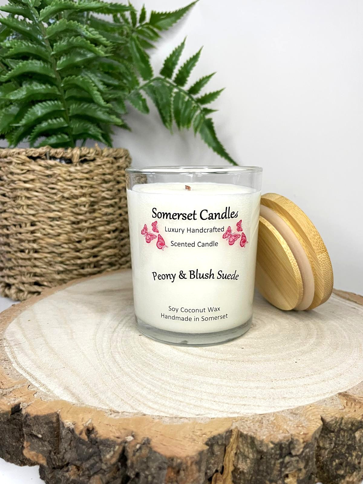 White soy candle in a 7oz clear glass jar with a natural wooden lid. The candle is made from a soy-coconut blend and scented with a soft elegant mix of Peony and Blush Suede. Above the scent name on the label, several pink butterflies are displayed, adding a romantic touch. Clean, minimalist design ideal for gifts, home decor or self-care.