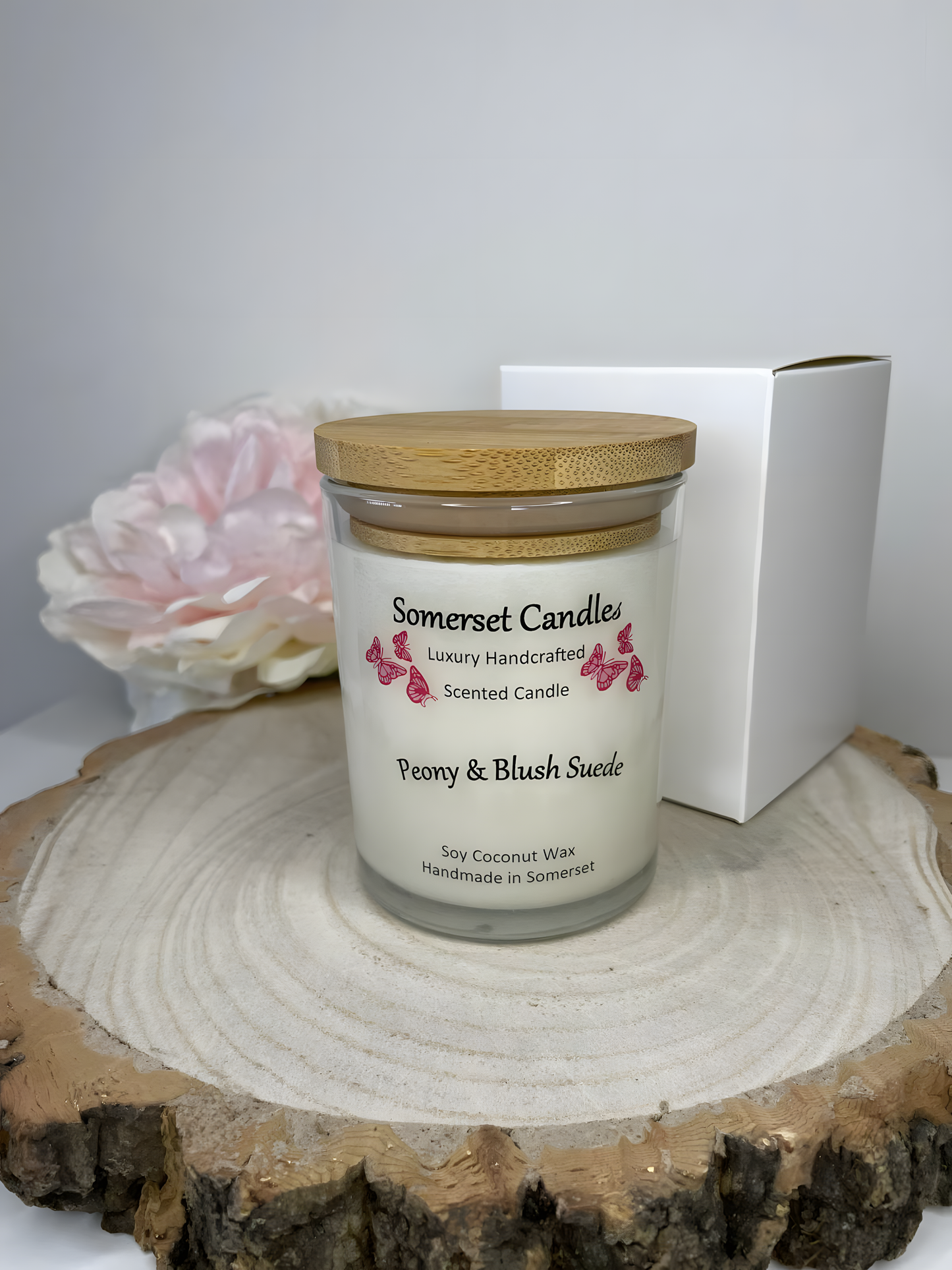 Peony & Blush scented soy candle suede displayed on a wooden board with gift box and a beautiful pink peony in the back ground. The candle is white in colour with bright pink butterflies fluttering above the scent name and fitted with a bamboo lid.