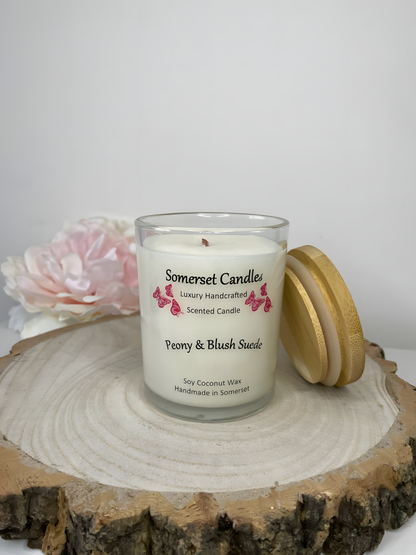 Peony & Blush scented soy candle suede displayed on a wooden board with a beautiful pink peony in the back ground. The candle is white in colour with bright pink butterflies fluttering above the scent name. A bamboo lid is leaning against the candle.