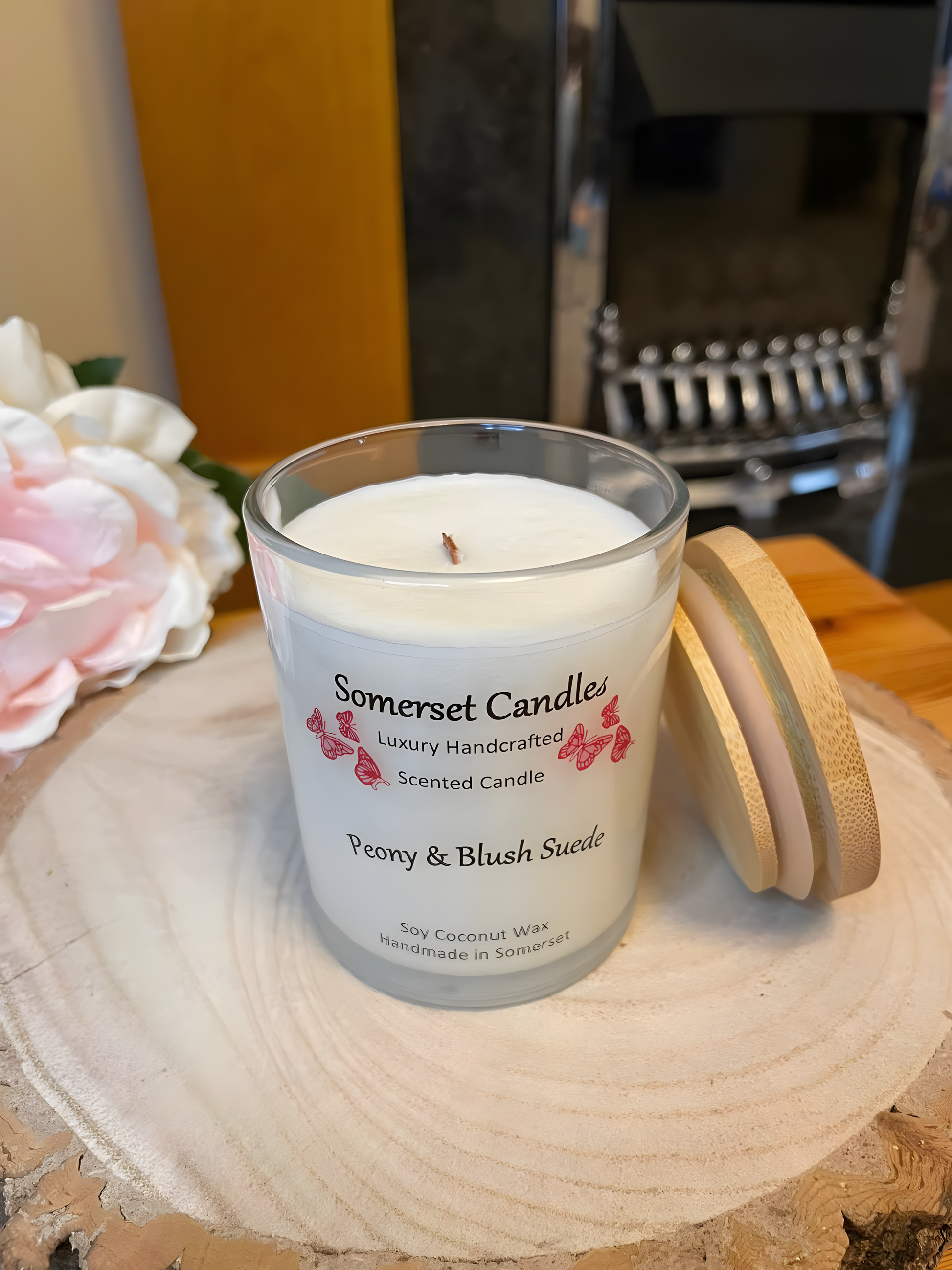 Peony & Blush scented soy candle suede displayed on a wooden board with a beautiful pink peony behind. The candle is white in colour with bright pink butterflies fluttering above the scent name. There&#39;s a bamboo lid lent against the candle.