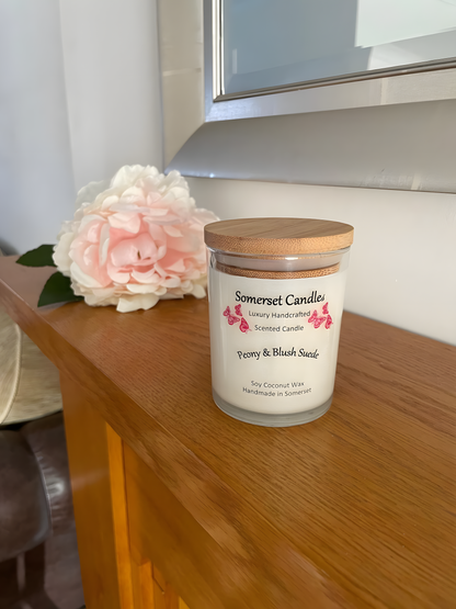 Peony & Blush scented soy candle suede displayed on a wooden mantel piece with a beautiful pink peony in the back ground. The candle is white in colour with bright pink butterflies fluttering above the scent name and fitted with a lovely bamboo lid.