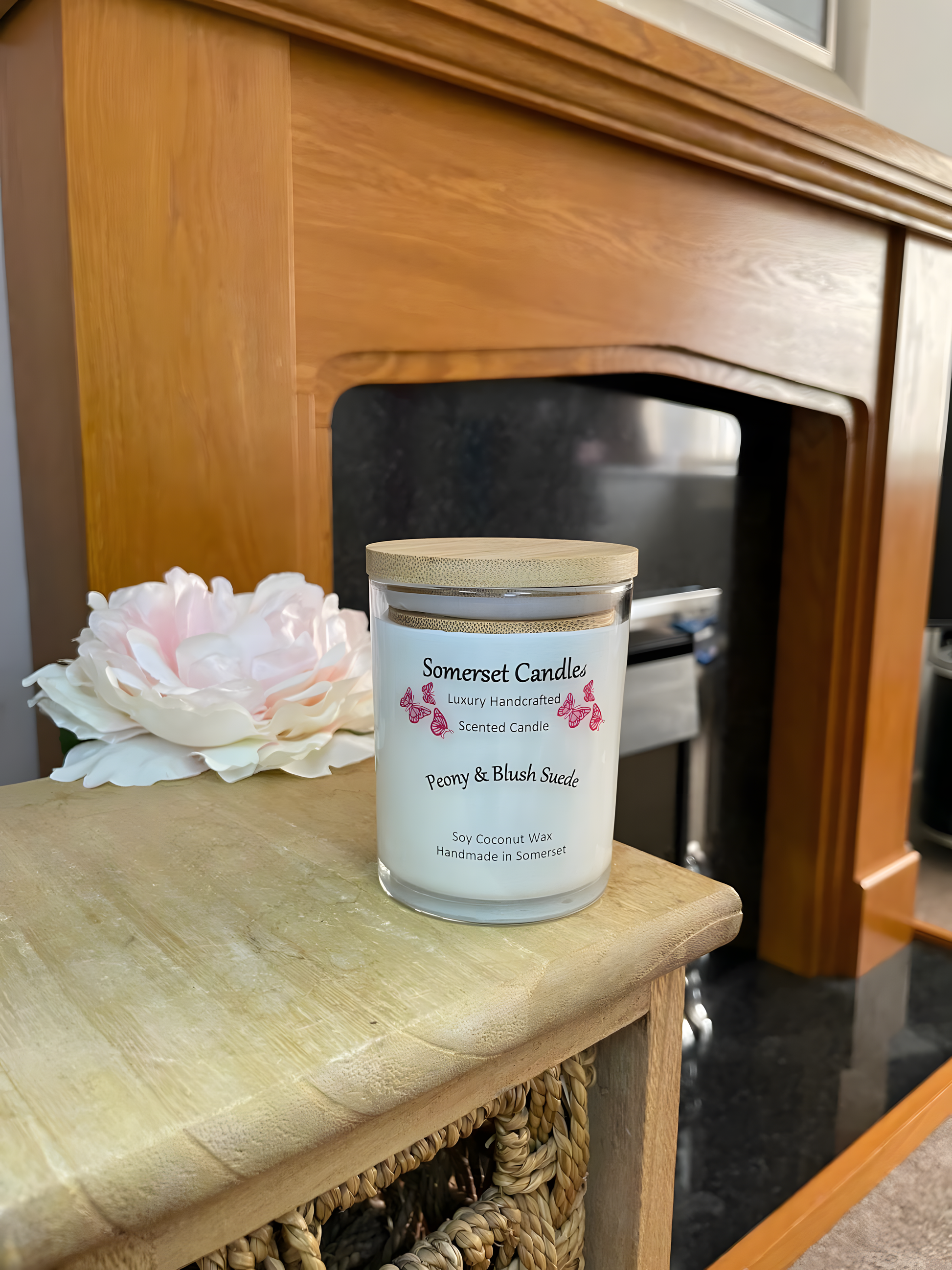 Peony & Blush scented soy candle suede displayed on a wooden board with fire and a beautiful pink peony in the back ground. The candle is white in colour with bright pink butterflies fluttering above the scent name and fitted with a lovely bamboo lid