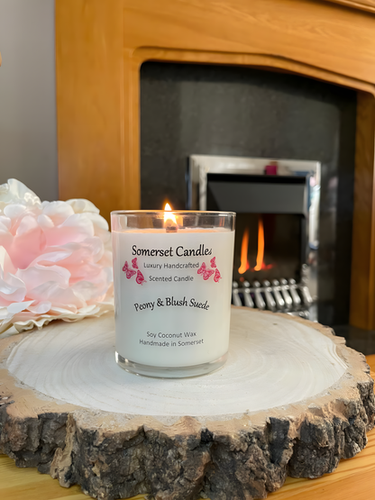 Peony & Blush scented soy candle suede displayed on a wooden board with a beautiful pink peony and a lit fire in the back ground. The candle is white in colour with bright pink butterflies fluttering above the scent name. The candle is lit.