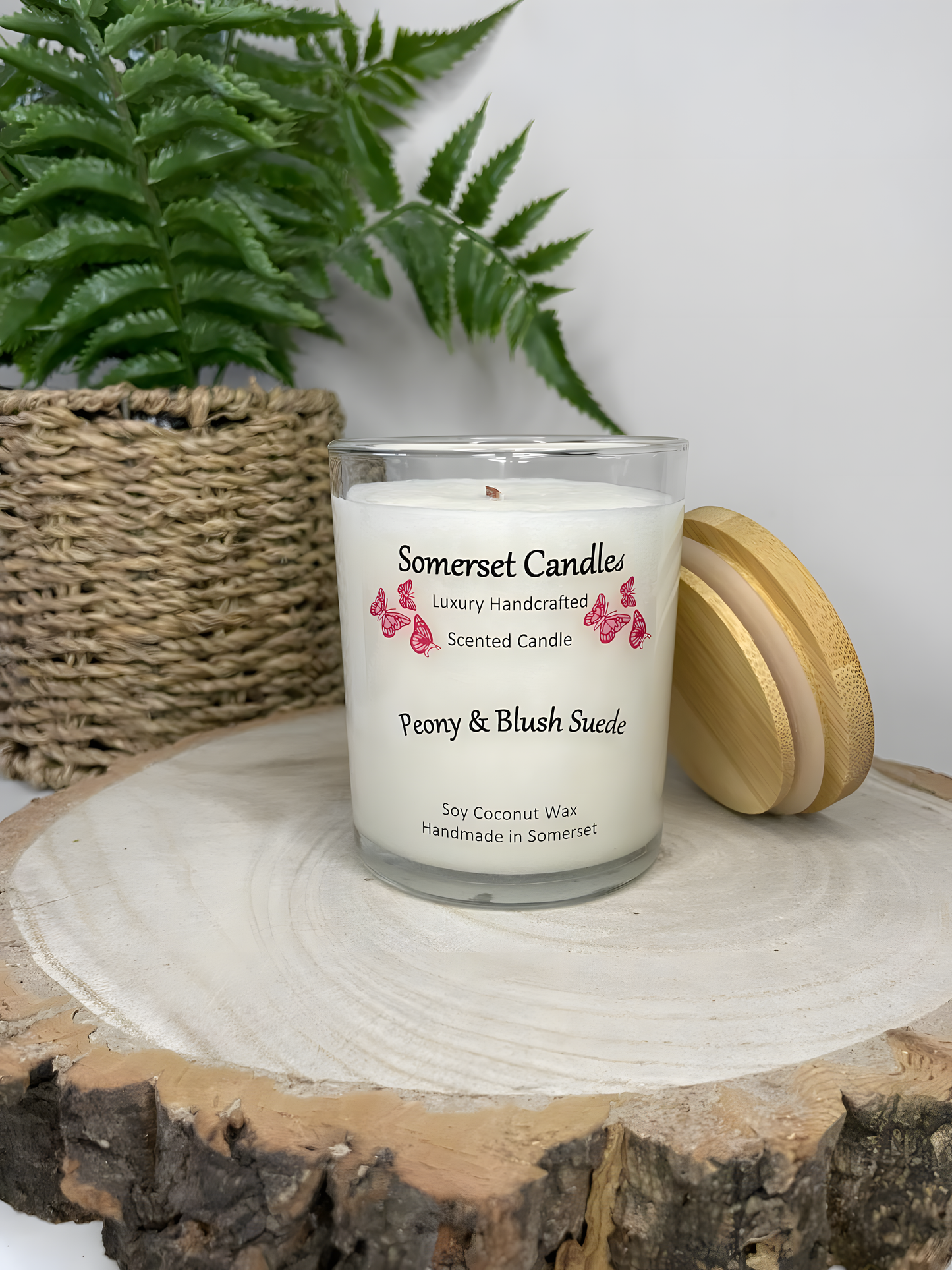 Peony & Blush scented soy candle suede displayed on a wooden board. The candle is white in colour with bright pink butterflies fluttering above the scent name. There is a lovely bamboo lid lent against the right side of the glass candle container.