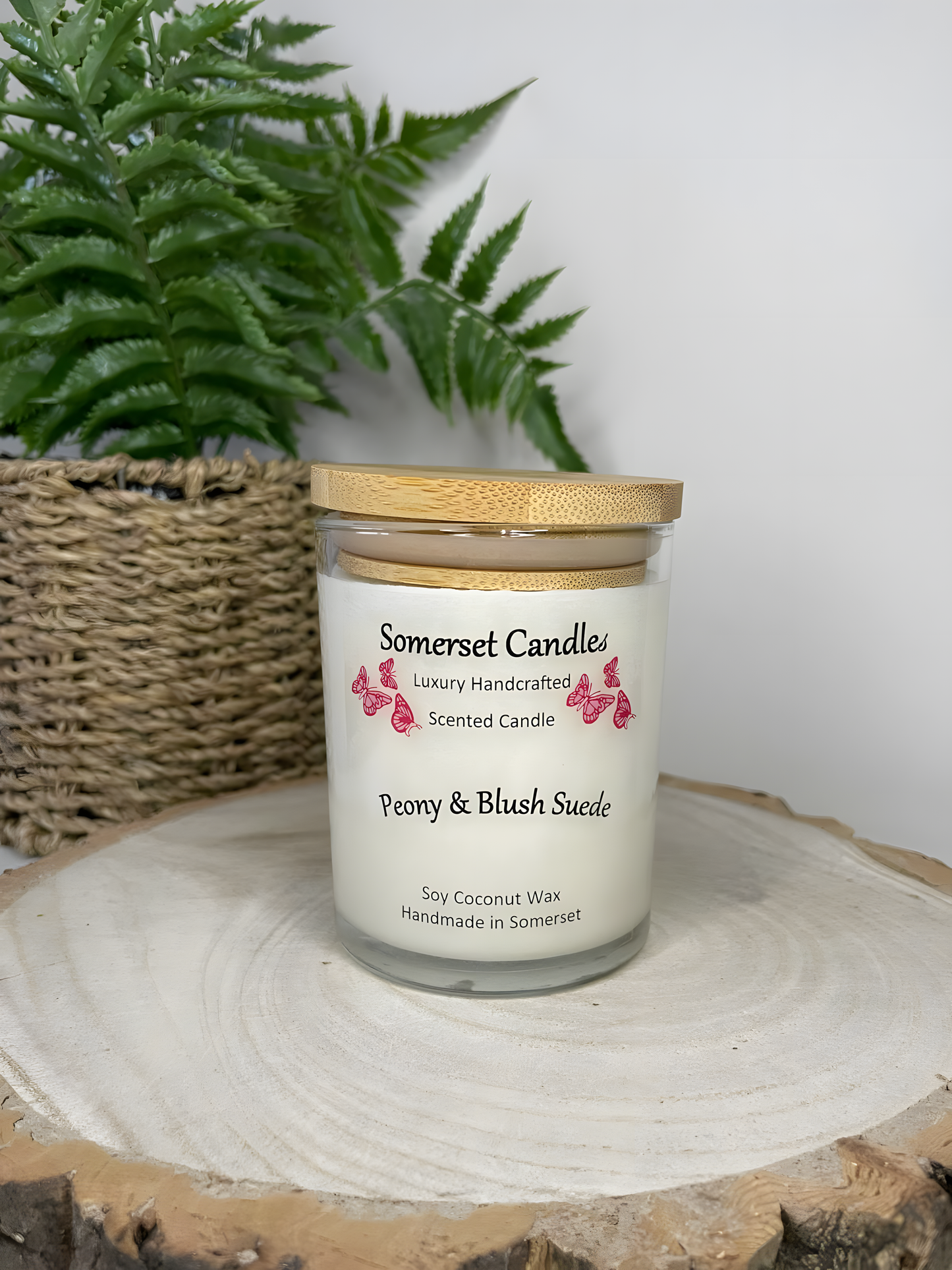 Peony & Blush scented soy candle suede displayed on a wooden board with a plant in the back ground. The candle is white in colour with bright pink butterflies fluttering above the scent name and fitted with a lovely bamboo lid.