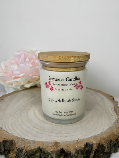 Peony & Blush scented soy candle suede displayed on a wooden board with a beautiful pink peony in the back ground. The candle is white in colour with bright pink butterflies fluttering above the scent name and fitted with a lovely bamboo lid.